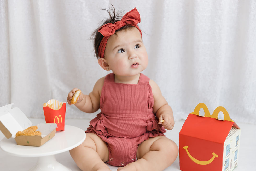Baby in a red outfit with headband holds a nugget, sitting beside fast food packaging on white table. White curtain backdrop. Curious expression.