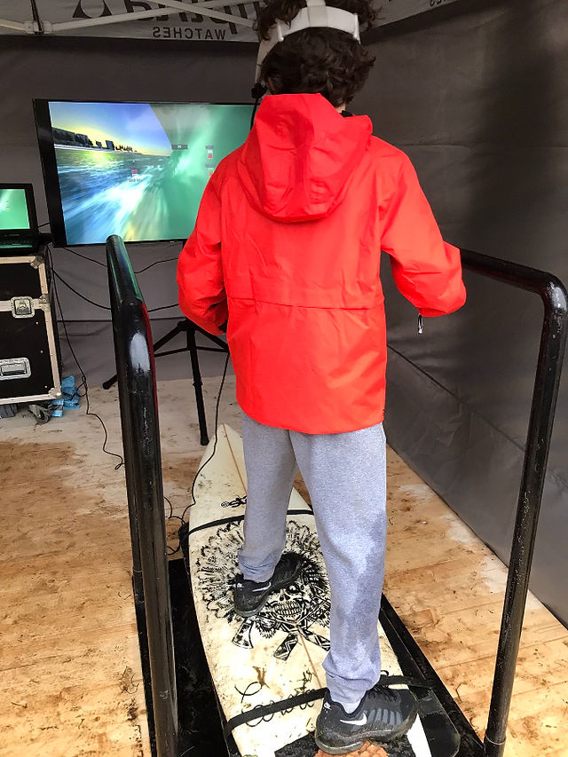 A boy in a red jacket is on a surf board attached to a unit in front of a big TV screen. In virtual reality he is surfing.