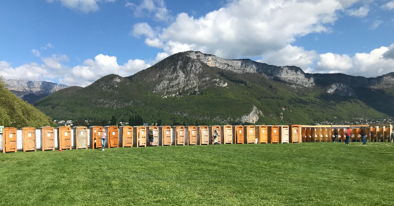 A row of twenty or more wooden portaloos are lined up on the Paquier, Annecy with the mountains as a backdrop. 