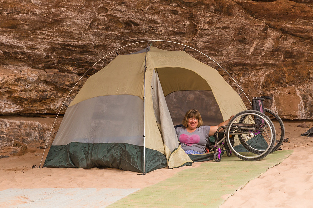 A woman sits inside a tent while extending her arm to her wheelchair outside of the tent.