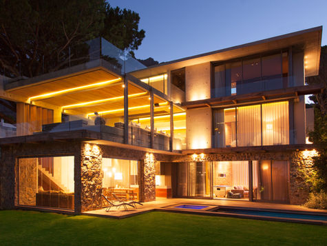 Modern luxury home illuminated at twilight, showcasing its sleek glass-and-stone architecture and a cozy, well-lit interior that opens to a lush, manicured garden.