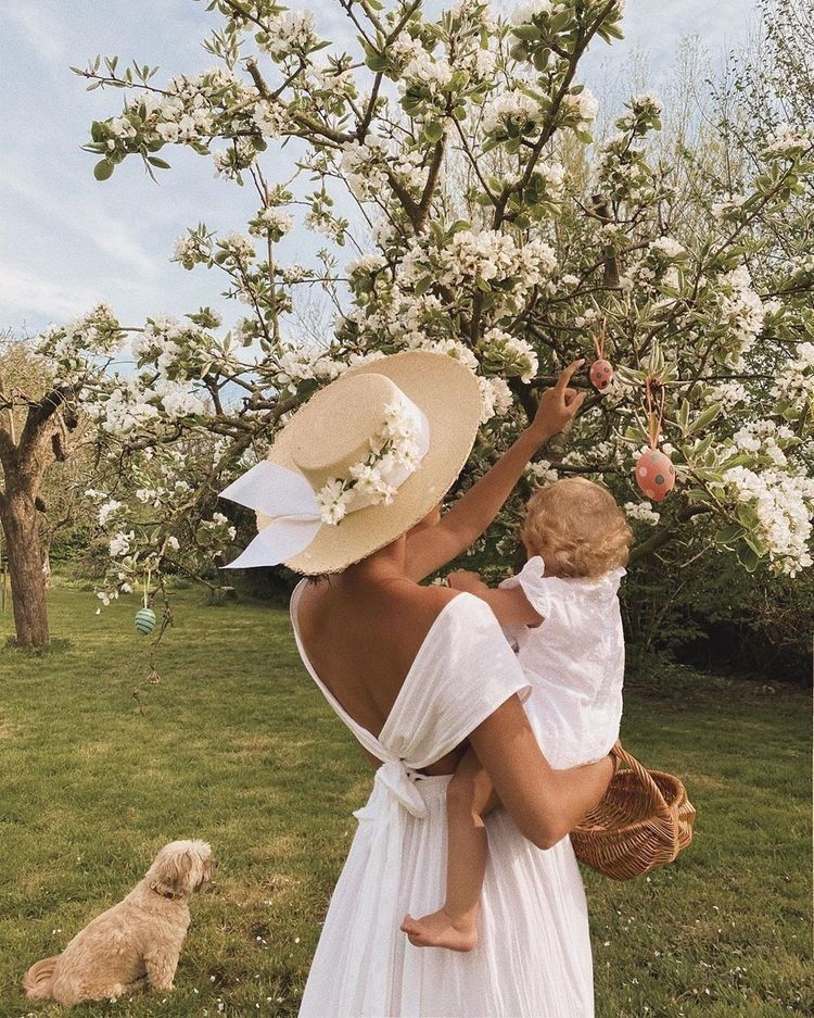 Mother in a white dress holding her child under a blooming tree in a sunlit garden, with a small dog nearby—celebrating family, nature, and safe, healthy living.