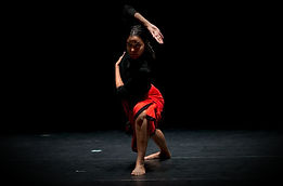 A contemporary dancer performs a grounded pose influenced by Payatt movement under a spotlight on a dark stage.