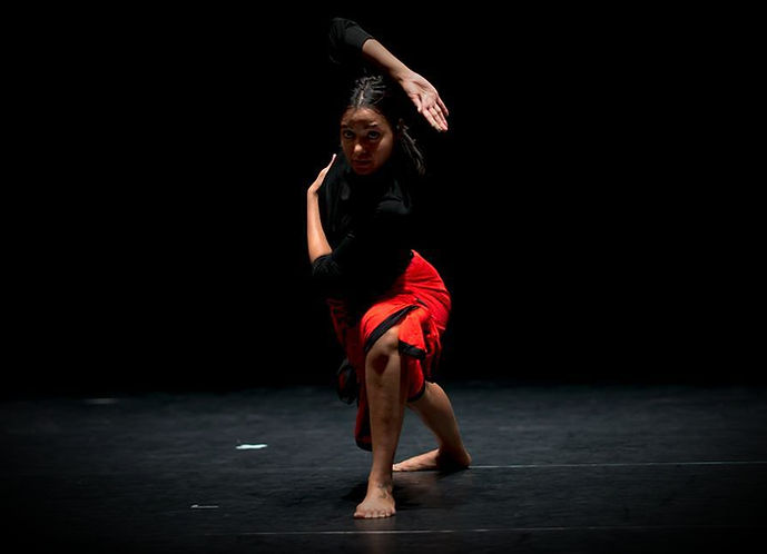 A contemporary dancer performs a grounded pose influenced by Payatt movement under a spotlight on a dark stage.