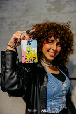 משתתפת מחייכת באירוע OPTICANA ✦ Offsite מציגה למצלמה את תג השם (Lanyard) הממותג שלה. התג מעוצב בשפת הפופ-ארט הססגונית של האירוע, הכוללת איורי דמויות עם משקפי שמש והכיתוב "OPTICANA". המשתתפת לבושה בסטייל מודרני המשתלב עם רוח המותג. הפקה: חגי מגידו – HM Productions.