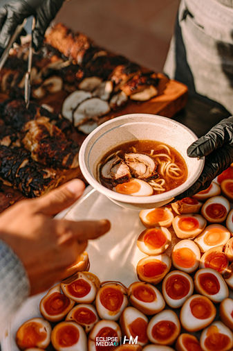 תצלום תקריב של עמדת הגשת אוכל אסייתי פרימיום באירוע Evoke ✦ LET LIGHT SHINE. נראית יד בכפפה שחורה מגישה קערת ראמן חמה עם אטריות וביצה חצי קשה, על רקע מגש עמוס בחצאי ביצים בעלות חלמון כתום עז ונתחי בשר מעושן על לוחות עץ. הפקה: חגי מגידו – HM Productions.