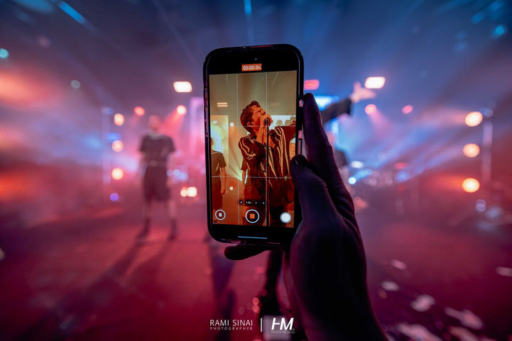תקריב של יד אוחזת במכשיר סמארטפון המתעד בוידאו את המופע על הבמה באירוע Illusion ✦ Transmit. על מסך הטלפון נראה הפרפורמר בבירור, בעוד הרקע האמיתי מטושטש באפקט בוקה עם אורות במה ורודים וכחולים. צילום: רמי סיני | הפקה: חגי מגידו – HM Productions.