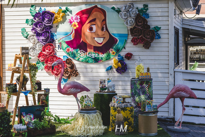 עמדת צילום (Photo Op) מעוצבת ומרהיבה באירוע LOGZ ✦ TIKI CHIC. במרכז הקיר מופיע איור פופ-ארט עגול וגדול של דמות נשית בווייב טרופי עם לוגו חברת Logz.io, מוקף בפרחי נייר ענקיים בעבודת יד, פסלי פלמינגו ורודים, חצאיות קש דקורטיביות ואלמנטים של צמחייה טרופית. הפקה: חגי מגידו – HM Productions.