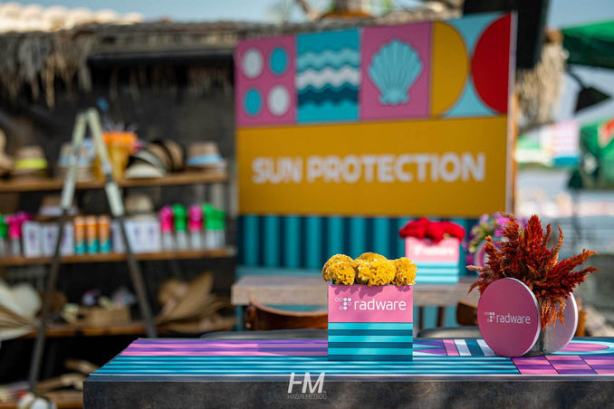 תקריב על עמדת הגנה מהשמש באירוע Summer Festival ✦ Radware. בחזית נראה שולחן ממותג בפסים כחולים וורודים עם סידורי פרחים צהובים וארגמניים בתוך כלים הנושאים את לוגו Radware. ברקע ניצב שלט צהוב גדול עם הכיתוב "SUN PROTECTION" ודפוסים של צדפים וגלים, ולצידו מדפים עם כובעי קש ומוצרי הגנה. הפקה: חגי מגידו – HM Productions.
