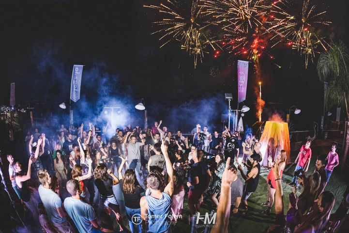 מבט פנורמי לילי על רחבת הריקודים העמוסה באירוע LOGZ ✦ TIKI CHIC. מאות עובדי Logz.io חוגגים תחת שמיים המוארים במופע זיקוקי דינור מרהיב, כשברקע נראים אלמנטים של תפאורת הטיקי הכוללים "הר געש" מואר ואפקטים של עשן וערפל המעצימים את האווירה החגיגית. הפקה: חגי מגידו – HM Productions.