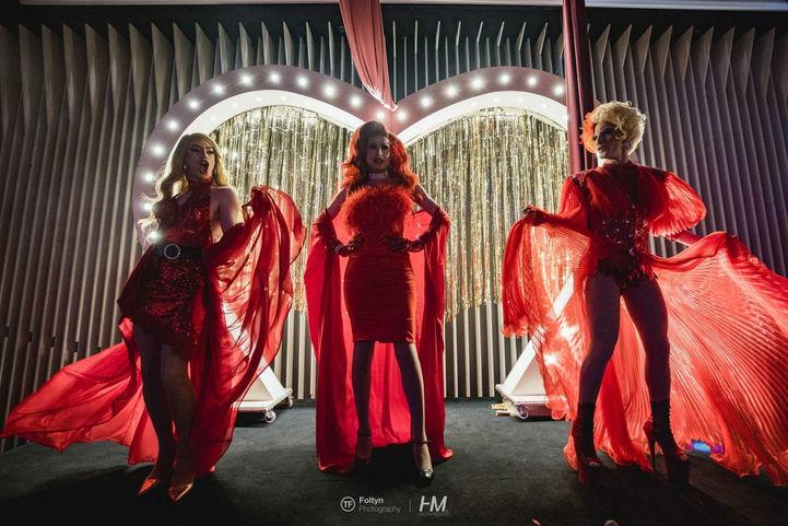 שלוש פרפורמריות בלבוש אדום דרמטי עם גלימות מרשימות עומדות בפוזות תיאטרליות על הבמה באירוע Molan Rouge ✦ Kali. ברקע נראה מיצג הלב המואר והנוצץ המשלים את האווירה היוקרתית. הפקה: חגי מגידו – HM Productions.
