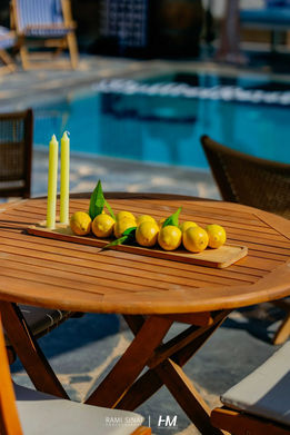 שולחן עץ עגול על שפת בריכה באירוע ShutterResort ✦ Shutterfly. על השולחן מגש עץ מאורך עם שורת לימונים צהובים ושני נרות דקים בגוון לימון. הפקה ועיצוב: חגי מגידו – HM Productions