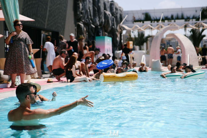 משתתפים נהנים מזמן איכות בבריכה ומסביבה באירוע Evoke ✦ ALOHAA. ברקע נראים עובדים משוחחים על שפת הבריכה, בעוד אחרים שוחים ומשחקים עם אביזרים מתנפחים ומטקות במים התכולים. המתחם מעוצב עם אלמנטים פיסוליים ורודים ופינות ישיבה מוצלות, תחת אור יום בהיר. הפקה: חגי מגידו – HM Productions.