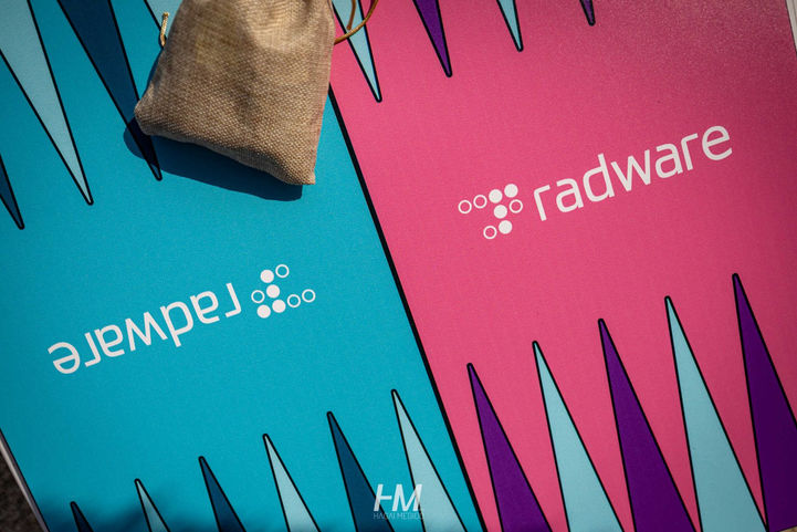 תקריב מלמעלה על לוח שש-בש מעוצב וממותג עבור אירוע Summer Festival ✦ Radware. הלוח מחולק לשני צדדים, ורוד ותכלת, עם הלוגו הלבן של חברת Radware במרכז כל צד. בפינה מונח שקיק בד קטן המכיל את כלי המשחק, על רקע הצורות הגיאומטריות של הלוח. הפקה: חגי מגידו – HM Productions.