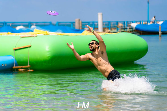 משתתף באירוע Drive-net ✦ Club קופץ לתוך המים בניסיון לתפוס צלחת מעופפת (פריזבי) סגולה, כשברקע נראה מתקן מתנפח ירוק ענק על פני הים. התמונה לוכדת רגע של אקשן, צחוק ופעילות גופנית. הפקה: חגי מגידו – HM Productions.