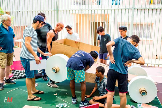 קבוצת משתתפים, מבוגרים ונוער, עובדים יחד בשיתוף פעולה על הרכבה ובנייה של מכונית מרוץ מקרטון עם גלגלים לבנים גדולים, כחלק מפעילות גיבוש חווייתית ותחרותית באירוע Atera ✦ Summer.
