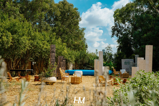 מבט רחב ומזמין על מתחם הלאונג' המרכזי באירוע TRANSMIT ✦ MOJO בשעות היום. המרחב כולל פינות ישיבה מראטן, עמודי תפאורה לבנים גבוהים ובריכה כחולה בולטת ברקע, כולם משולבים בתוך חורשה ירוקה ועבותה תחת שמיים בהירים. הפקה: חגי מגידו – HM Productions.