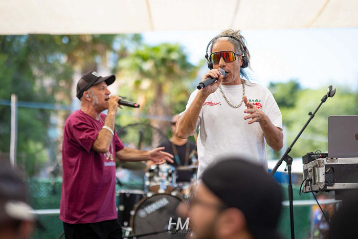 הופעה חיה על הבמה באירוע Summer Festival ✦ Radware. שני סולנים שרים בלהט לתוך מיקרופונים, אחד בחולצה בורדו והשני בחולצה לבנה עם משקפי שמש אופנתיים, על רקע מערכת תופים וצמחייה ירוקה תחת צילייה לבנה גדולה. הפקה: חגי מגידו – HM Productions.