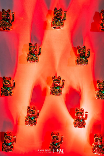 תקריב אמנותי של קיר דקורטיבי המורכב מעשרות פסלוני "Maneki-neko" (חתול המזל היפני) מסודרים בסימטריה מושלמת. הקיר כולו שטוף בתאורה אדומה עזה ודרמטית, המדגישה את צלליות החתולים המניפים את כפותיהם כסמל להזמנת מזל, שפע והצלחה. המקצב הוויזואלי החוזר מייצר אפקט של קיר כוח מרשים בלב האירוע. הפקה: חגי מגידו – HM Productions.