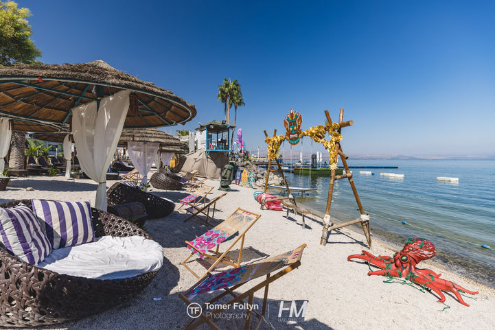 מבט רחב על רצועת החוף המעוצבת באירוע LOGZ ✦ TIKI CHIC. המתחם כולל שמשיות קש רחבות עם וילונות לבנים מתנפנפים, מיטות שיזוף יוקרתיות מקלועי ראטן, וכיסאות נוח ממותגים בהתאמה אישית. ברקע ניצבת נדנדת הבמבוק האייקונית עם מסיכת הטיקי, ולצדה פסל תמנון אדום גדול על החול, אל מול המים הצלולים והשמיים הכחולים. הפקה: חגי מגידו – HM Productions.