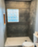 Shower under renovation with gray paneled walls, frosted glass block window, and white toilet. Roll of toilet paper on the tank.