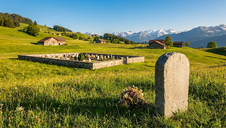 Auf einer grünen Wiese in einer idyllischen Berglandschaft liegt ein kleiner Friedhof mit schlichten Grabsteinen, umgeben von traditionellen Holzhäusern und majestätischen Alpen. Ein einsamer Gedenkstein und ein Blumenstrauß betonen die stille Schönheit der Szene.