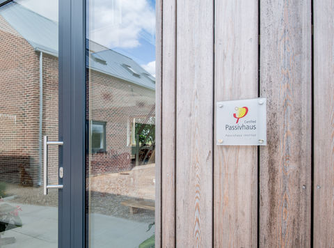 Certified Passivhaus sign stands proud of wood cladding next to glazed door with reflection of other parts of the new build in view