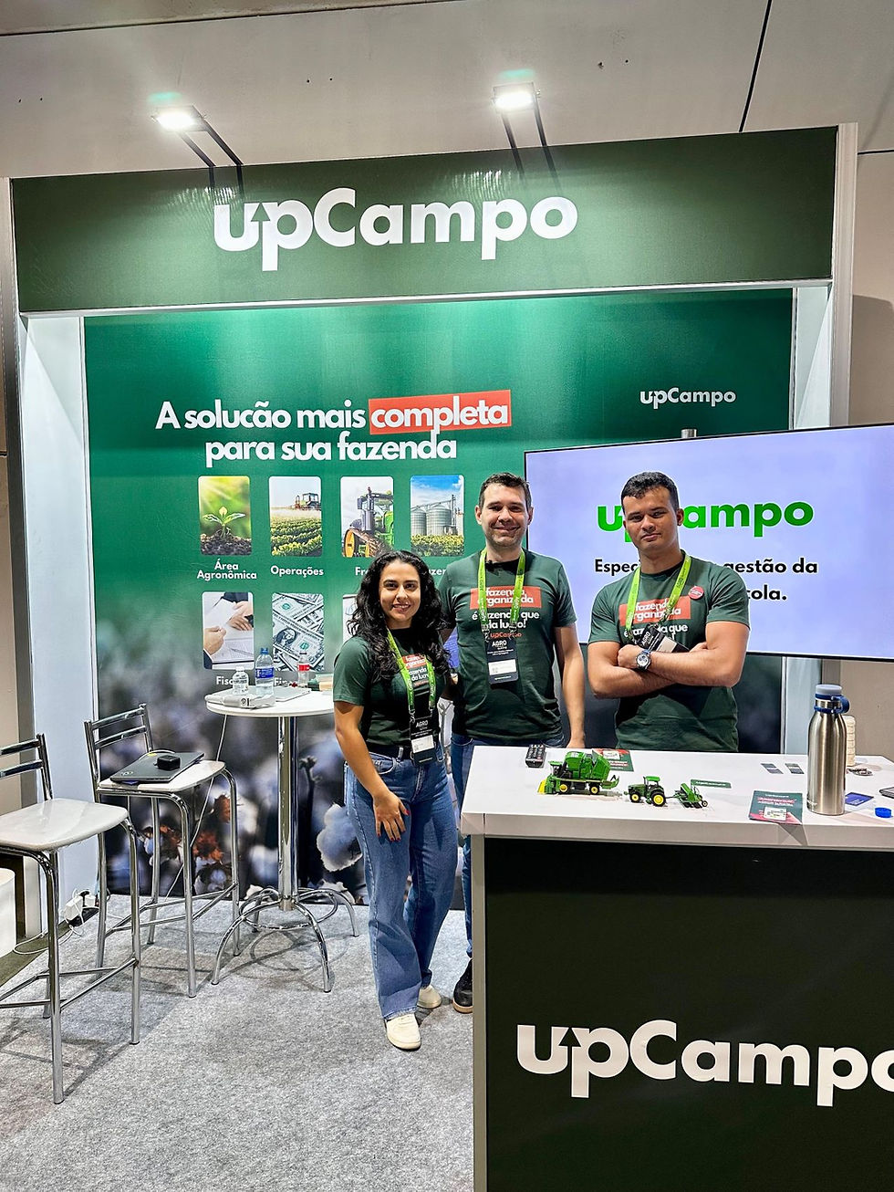 Três pessoas sorridentes em um estande da UpCampo, com fundo verde e textos promocionais. Há miniaturas de tratores e clima descontraído.