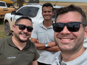 Três homens sorriem em um campo com colhedora amarela e caminhonetes ao fundo. Céu azul com nuvens. Camisetas da empresa upCampo.