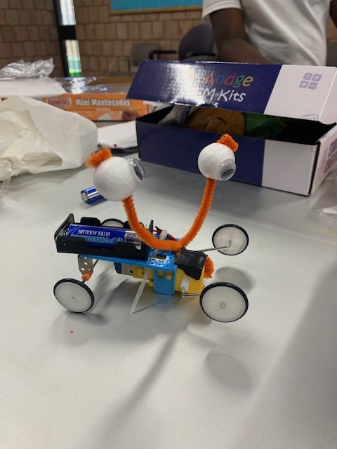 A handmade wheeled robot decorated with googly eyes and orange pipe cleaners on a classroom table