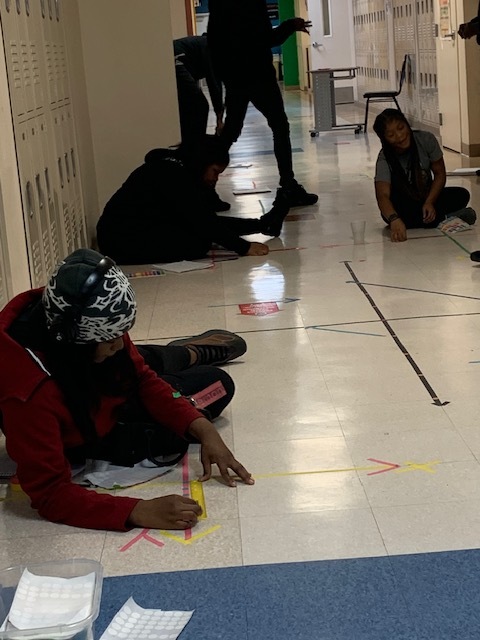 Students working with taped geometric lines and measuring tools on a school hallway floor