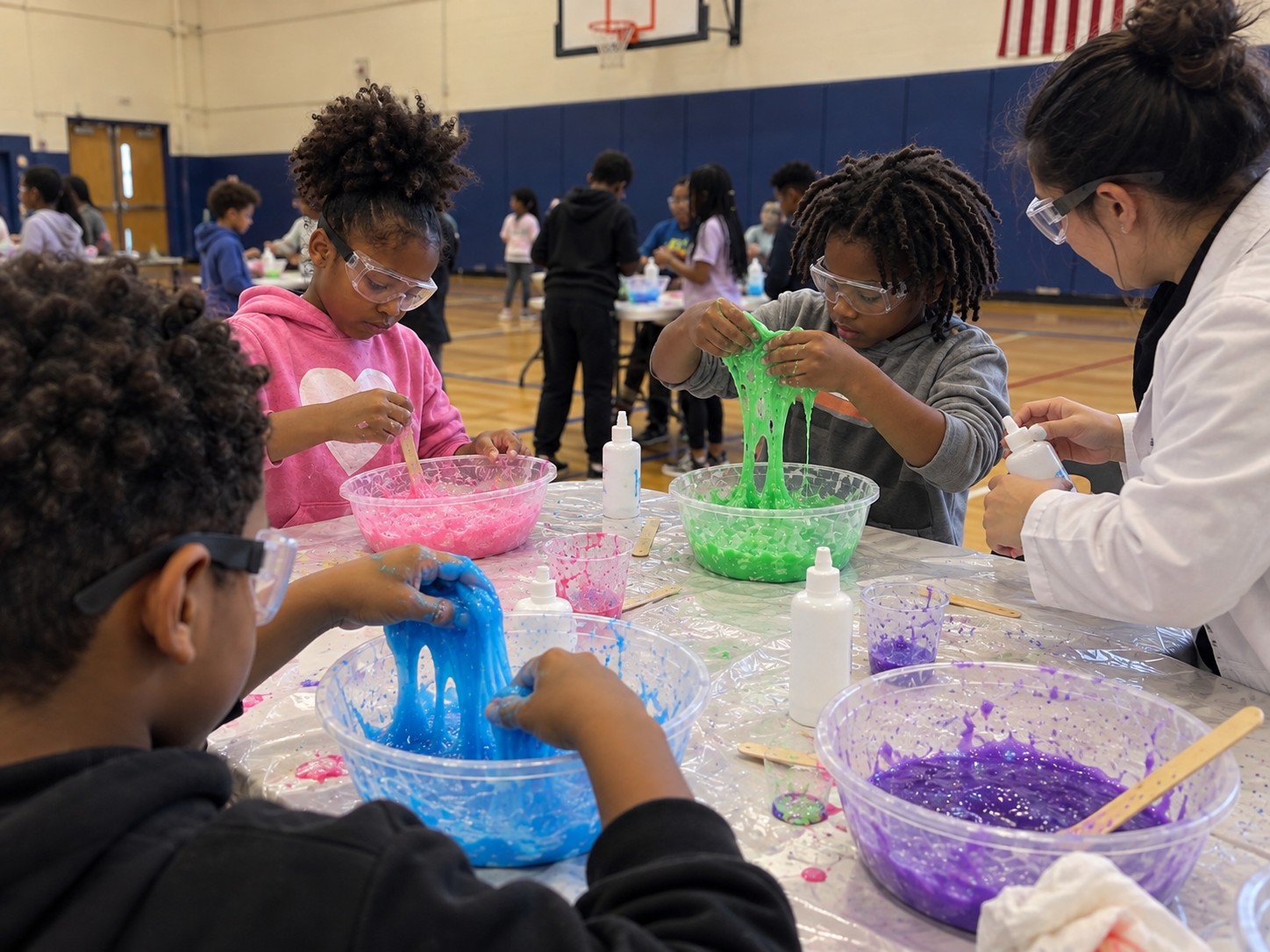 Students wearing clear safety goggles, mixing and stretching colourful slime at a table in a school gym