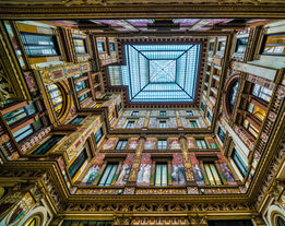 Looking up through the interior courtyard of Palazzo Braschi in Rome, Italy, featuring ornate architectural details, colorful facades, symmetrical balconies, and a glass skylight overhead.