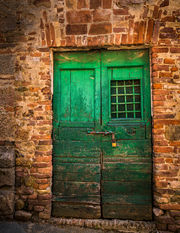 A weathered green wooden door with an iron-grilled window set in a rustic stone wall in a medieval village in Tuscany, Italy, showcasing historic architecture and aged textures.