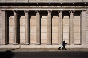 A lone pedestrian walks past the tall stone columns of a neoclassical building in the City of London financial district, highlighting symmetry, architecture, and urban design.