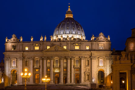 St. Peter's Basilica in Vatican City photographed at night, featuring its illuminated Renaissance façade, iconic dome, and glowing architectural details beneath a deep blue evening sky.