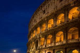 Close-up of the illuminated Colosseum in Rome, Italy, photographed at night, featuring glowing arches, ancient stone architecture, and a deep blue evening sky with the moon in the background.