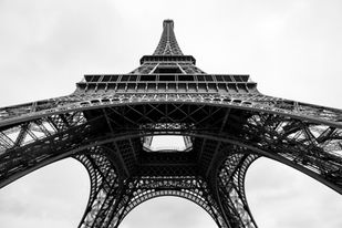 Black-and-white photograph looking up from beneath the Eiffel Tower in Paris, France, showcasing its intricate iron lattice structure, dramatic perspective, and iconic architectural design.