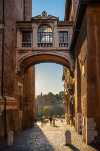 The Bridge of Sighs in Rome, Italy, spanning a narrow cobblestone street and framing a view toward the Tiber River with historic buildings bathed in warm morning light.