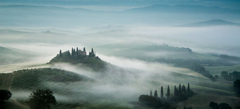 Morning fog blankets the rolling hills of Tuscany, Italy, surrounding a hilltop grove of cypress trees and creating a serene, misty landscape at sunrise.