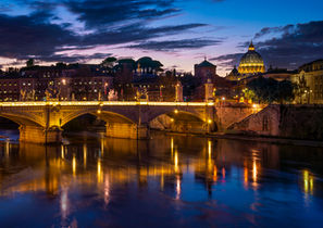 St. Peter's Basilica in Vatican City viewed from the Tiber River at twilight, featuring illuminated bridges, colorful reflections on the water, and a dramatic blue and purple evening sky.