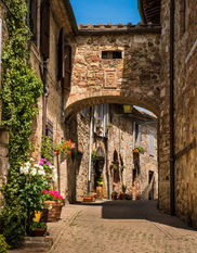 A narrow cobblestone street in a medieval village in Tuscany, Italy, featuring stone buildings, an elevated stone archway, colorful flower pots, and historic Italian architecture.