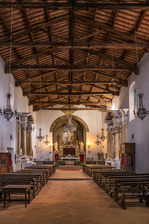 Interior of a historic chapel in Tuscany, Italy, featuring exposed wooden ceiling beams, rows of wooden pews, rustic stone walls, decorative lanterns, and a traditional altar.