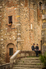 Two young priests sitting on stone steps beside a historic medieval building in a Tuscan village, Italy, surrounded by weathered stone architecture and a peaceful cobblestone street.