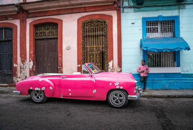 A bright pink vintage American convertible parked on a colorful street in Havana, Cuba, with historic colonial buildings and a local resident standing nearby.