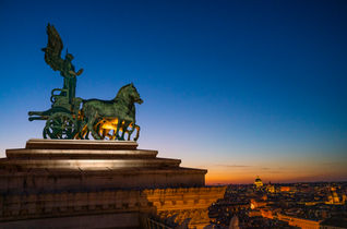 Bronze quadriga statue atop the Victor Emmanuel II Monument in Rome, Italy, silhouetted against a vibrant sunset with panoramic views of the city skyline.