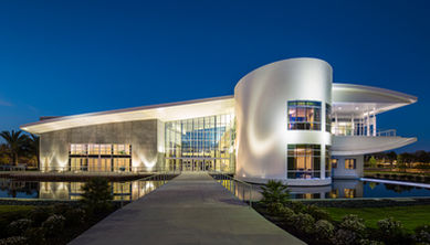 The Student Union building at Embry-Riddle Aeronautical University in Daytona Beach, Florida, photographed at dusk, featuring curved contemporary architecture, floor-to-ceiling glass, dramatic exterior lighting, and reflecting pools.