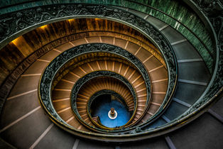Looking down into the Bramante Staircase at the Vatican Museums in Vatican City, featuring a double-helix spiral staircase, ornate iron railings, and striking geometric symmetry.