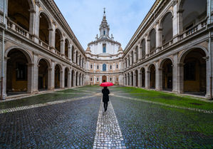 Courtyard of Sant'Ivo alla Sapienza in Rome, Italy, featuring symmetrical arcades, cobblestone walkways, and a person holding a red umbrella leading toward the historic Baroque church.