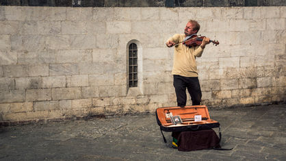 A street musician playing the violin in Siena, Tuscany, Italy, performing beside a historic stone wall with an open instrument case in the foreground.
